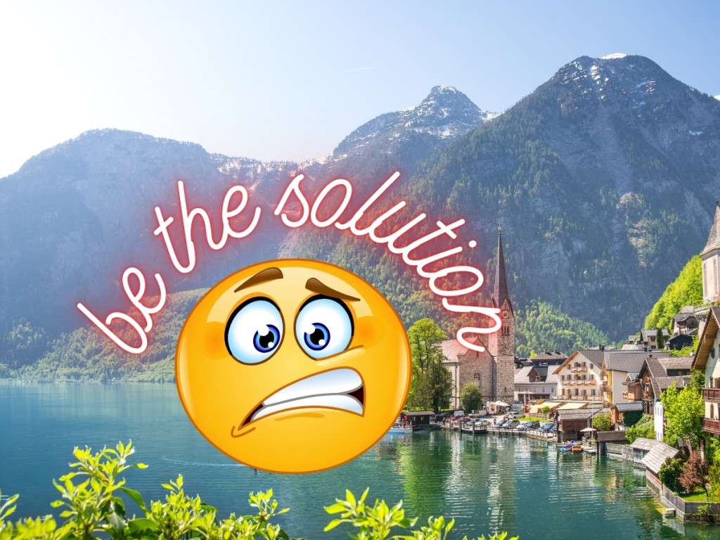 How to visit Hallstatt cover photo. the image depicts the famous town of Hallstatt Austria, a small town on the banks of a lake, with towering mountains in the background. In the foreground of the photo is a concerned looking emoji with the wording "be the solution"
